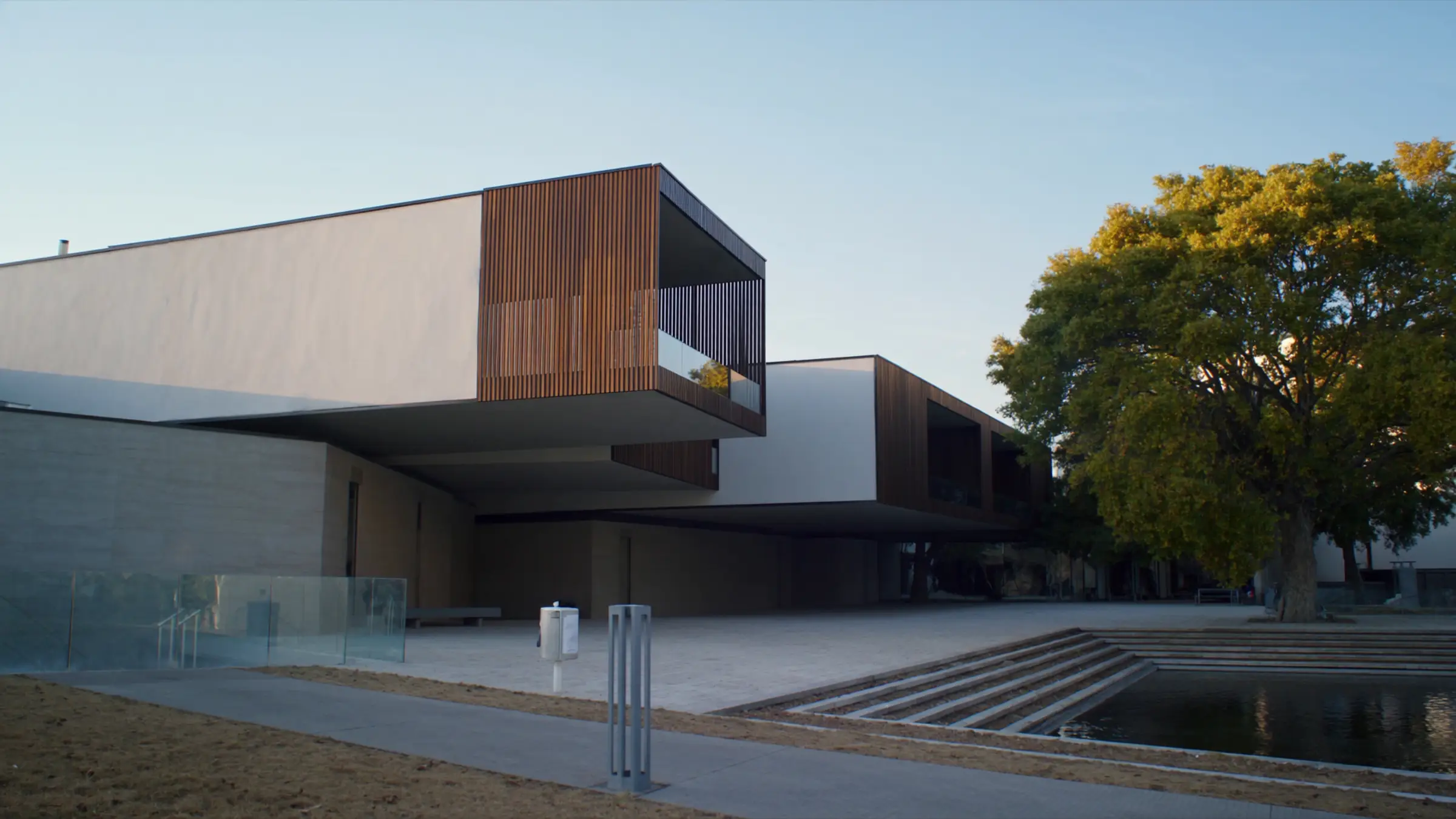 A modern low-rise building in white and timber, with a landscaped plaza and reflecting pool at sunset